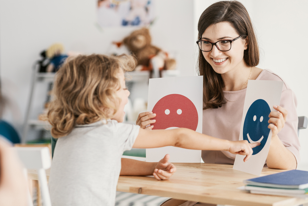 Logopeda prowadzi <strong>terapię logopedyczną</strong> z dzieckiem - terapeuta trzyma kartki z twarzami wyrażającymi nastroje